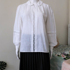 May include: A white long-sleeved blouse with a peter pan collar and lace trim. The blouse has a button-up front and a relaxed fit.