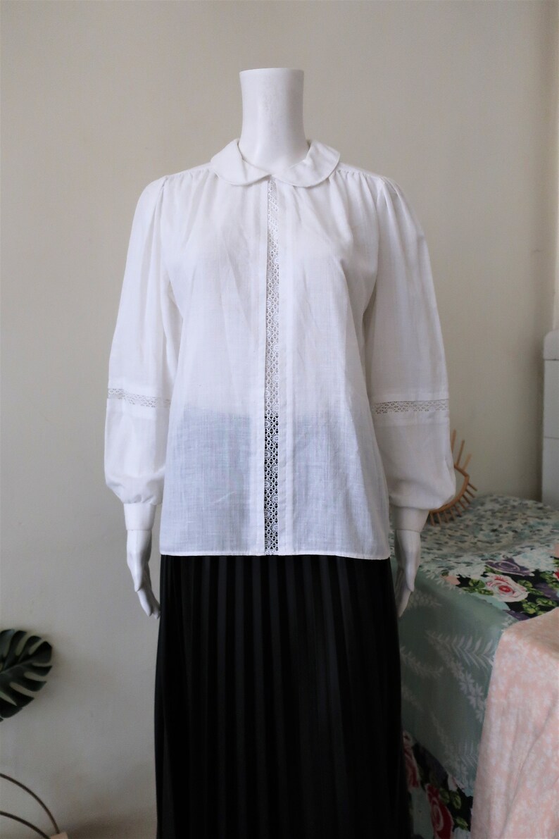 May include: A white long-sleeved blouse with a peter pan collar and lace trim. The blouse has a button-up front and a relaxed fit.