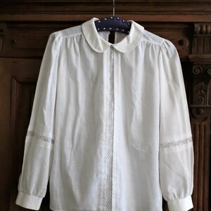 May include: A white long-sleeved blouse with a peter pan collar and lace trim on the sleeves and down the front.