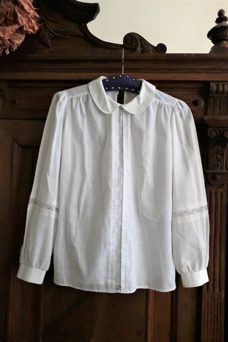 May include: A white long-sleeved blouse with a peter pan collar and lace trim on the sleeves and down the front.