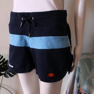 Puede incluir: Shorts deportivos azules y negros con una franja azul y un logotipo en la esquina inferior derecha. Los shorts tienen una cintura con cordón.