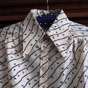 Puede incluir: Una camisa blanca de botones con un patrón geométrico azul y blanco. La camisa tiene un cuello puntiagudo y mangas largas.