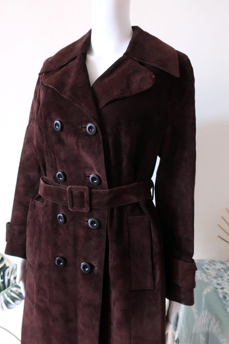 May include: A long brown suede trench coat with a double breasted closure, a belt, and a collar.
