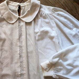 May include: A white cotton blouse with a peter pan collar and lace trim. The blouse has long sleeves with lace trim at the cuffs.