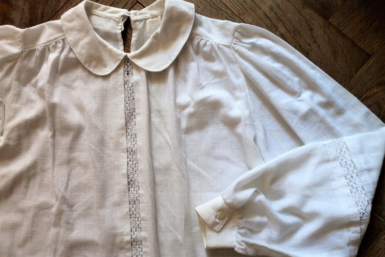 May include: A white cotton blouse with a peter pan collar and lace trim. The blouse has long sleeves with lace trim at the cuffs.