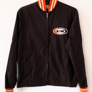 Op de afbeelding: Een bruine jas met ritssluiting en oranje en witte strepen op de kraag en manchetten. De jas heeft een A&W-logopatch op de borst. Hij hangt aan een metalen haak tegen een witte achtergrond.
