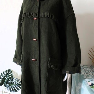 May include: Dark green hooded coat with fringe detailing across the chest and on the pockets. The coat has wooden buttons and plaid lining on the hood and cuffs. A stylish outerwear piece.