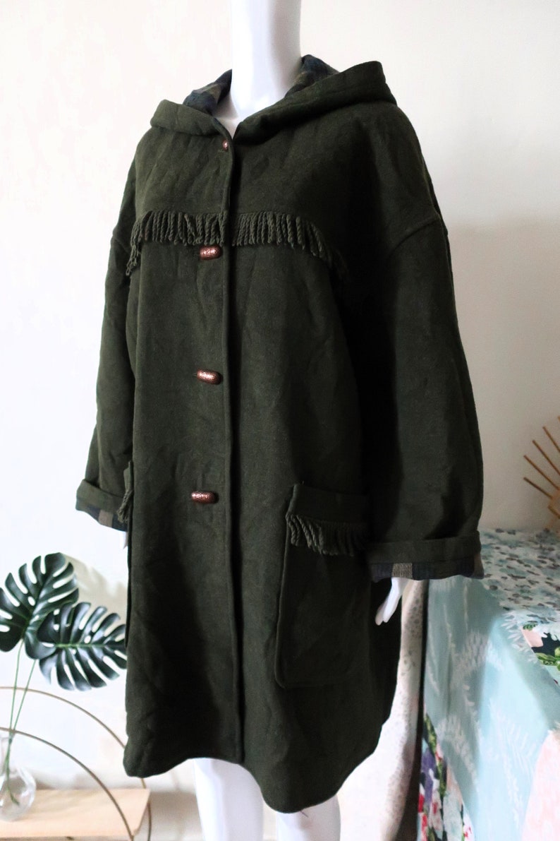 May include: Dark green hooded coat with fringe detailing across the chest and on the pockets. The coat has wooden buttons and plaid lining on the hood and cuffs. A stylish outerwear piece.