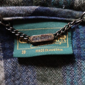 May include: Close-up of a wool jacket label with the brand name "Geiger" and "Made in Austria" printed on a green label. The jacket has a blue, green, and gray plaid pattern. A metal chain is attached to the label.