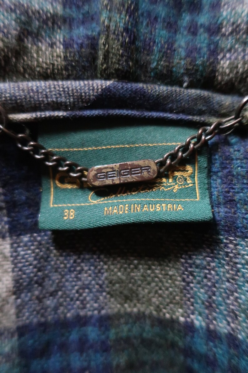 May include: Close-up of a wool jacket label with the brand name "Geiger" and "Made in Austria" printed on a green label. The jacket has a blue, green, and gray plaid pattern. A metal chain is attached to the label.