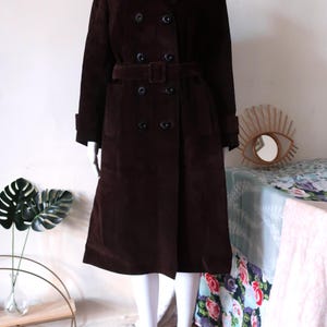 May include: A brown corduroy trench coat with a double breasted closure, a belt, and two pockets. The coat is on a white mannequin.