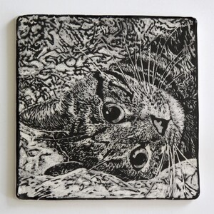 May include: Square black and white ceramic tile featuring a detailed illustration of a cat's face. The cat's large eyes and whiskers are prominent, with a textured background. The artwork is a close-up, artistic rendering of a feline.