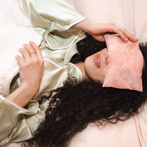 Op de afbeelding: Een persoon met lang, krullend donker haar ligt in bed met een roze oogmasker over haar ogen. Ze draagt een lichtgroen satijnen pyjama-top en een witte deken is tot aan haar borst getrokken.