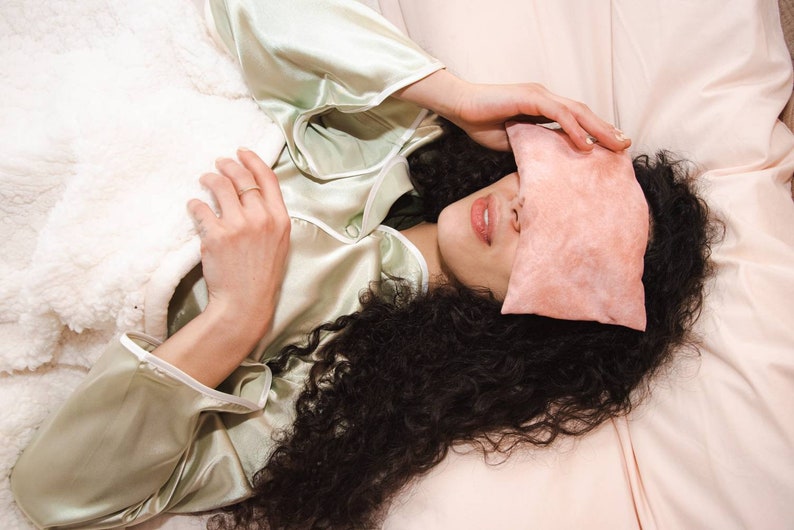 Op de afbeelding: Een persoon met lang, krullend donker haar ligt in bed met een roze oogmasker over haar ogen. Ze draagt een lichtgroen satijnen pyjama-top en een witte deken is tot aan haar borst getrokken.