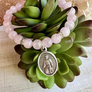 May include: A rose quartz beaded bracelet with a silver charm. The bracelet features round, light pink beads and a small, oval charm with an image of a saint and the inscription "ST. PEREGRINE". The bracelet is displayed on a green succulent plant.