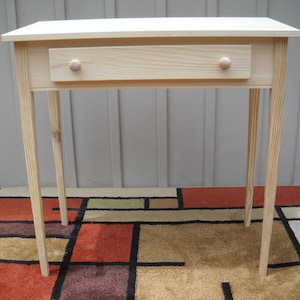 May include: A simple, unfinished wooden table with a single drawer and turned knobs. The table has four legs and is sitting on a patterned rug.