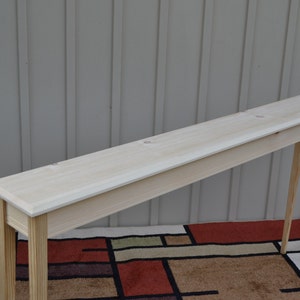 May include: A long, narrow, unfinished wooden console table with a light wood finish. The table has a rectangular top and four slender legs.