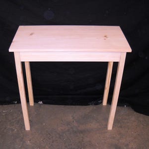 May include: A simple, unfinished wooden table with a rectangular top and four legs. The table is made of light-colored wood and has a natural, rustic look.