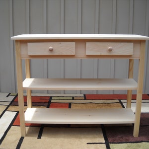 May include: A natural wood console table with two drawers and two shelves. The table is made of unfinished pine wood and has a simple, rustic design.