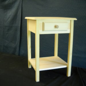 May include: A simple, unfinished wooden end table with a single drawer and a lower shelf. The table is made of light-colored wood and has a natural finish.