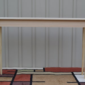 May include: A simple, light wood console table with a rectangular top and four slender legs. The table is standing on a patterned rug.