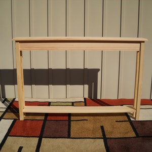 May include: A simple, unfinished wooden console table with a single shelf. The table is made of light-colored wood and has a rectangular shape. It is sitting on a patterned rug.