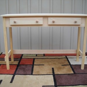 May include: A simple, unfinished wooden table with two drawers and a light wood finish. The table has a rectangular top and four legs. The table is sitting on a patterned rug.