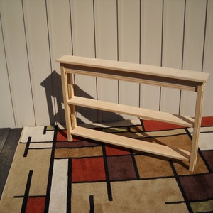 May include: A light-colored wooden console table with two shelves. The table is rectangular with a decorative top edge and is placed on a patterned rug. The table's dimensions are approximately 48 inches long, 12 inches wide, and 30 inches high.
