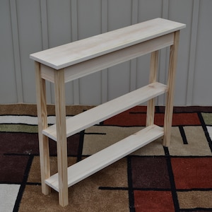 May include: A natural wood console table with two shelves. The table is made of unfinished pine and has a simple, rectangular design.