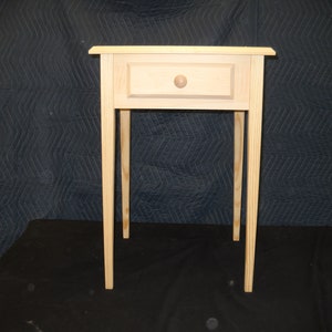 May include: A simple, wooden end table with a single drawer and turned legs. The table is made of unfinished pine wood.
