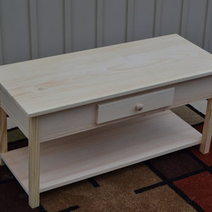 May include: A natural wood coffee table with a single drawer and a lower shelf. The table is unfinished and has a simple, rectangular design.