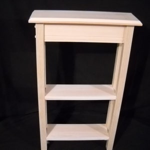 May include: A light-colored, unfinished wooden side table with three shelves. The table has a rectangular top and a simple, minimalist design. The shelves are evenly spaced, providing storage or display space.