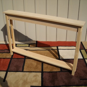 May include: A light wood console table with a simple design. The table has a rectangular top and a single shelf. The table is sitting on a patterned rug.