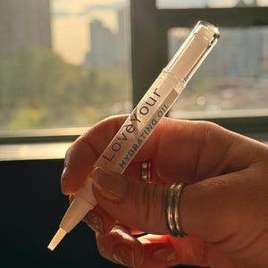 May include: A clear, pen-shaped hydrating oil applicator with the text "Love Your Hydrating Oil" in white and blue. The applicator is held in a hand with manicured nails and silver rings. The background shows a blurred cityscape.