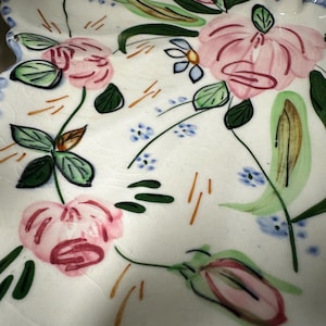 May include: A white ceramic serving dish with a scalloped edge, adorned with hand-painted pink flowers, green leaves, and small blue floral accents. The design includes delicate stems and orange brushstrokes, creating a charming, floral-themed piece.