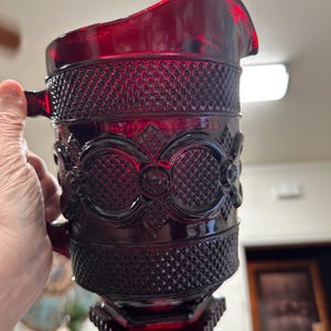 May include: A ruby red glass pitcher with a textured diamond pattern and ornate floral designs. The pitcher has a handle and a spout for pouring. The base is a faceted shape. The pitcher is held by a hand.