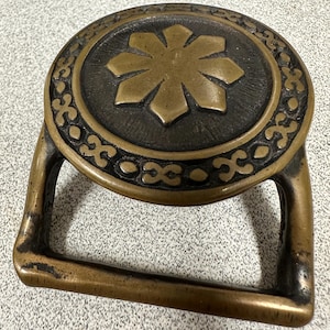 May include: A brass buckle with a decorative design. The buckle has a circular shape with a raised, star-shaped design in the center. The outer edge of the buckle is decorated with a repeating pattern.