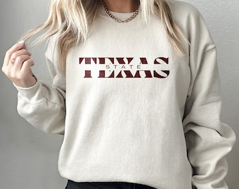 May include: A beige crewneck sweatshirt featuring the dark red text TEXAS with the word STATE printed in smaller letters centered between the T and the E. The garment has long sleeves and a ribbed neckline, cuffs, and hem, providing a relaxed fit.