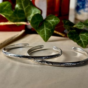 May include: Three silver-colored cuff bracelets with etched floral and leaf patterns. The bracelets are open-ended and rest on a neutral-toned surface. Green ivy leaves and amber-colored glass bottles are in the background.