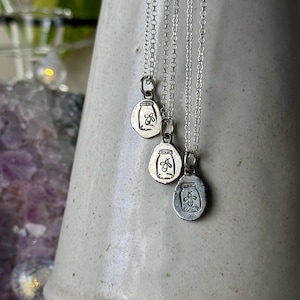 May include: Three silver necklaces with oval pendants. Each pendant features a jar design with a firefly inside. The necklaces hang in front of a white ceramic vase. The pendants are approximately 2 cm in length.