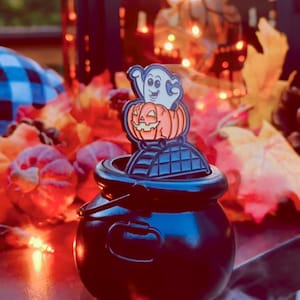 May include: A black metal cauldron with a handle, featuring a decorative enamel pin attached to the rim. The pin depicts a white ghost with a smiling expression sitting inside an orange jack-o'-lantern, which rests on a dark arched structure.