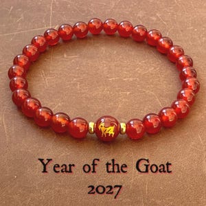 May include: A circular bracelet made of polished red beads with a central bead featuring a gold-coloured goat icon, flanked by two small gold-coloured spacers. Below the bracelet, the text reads Year of the Goat 2027 in a dark font.