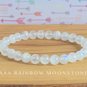 May include: A stretch bracelet composed of round, translucent white moonstone beads with subtle blue iridescence. The beads are strung in a single circular row. The text AAA RAINBOW MOONSTONE is printed in white capital letters below the bracelet.