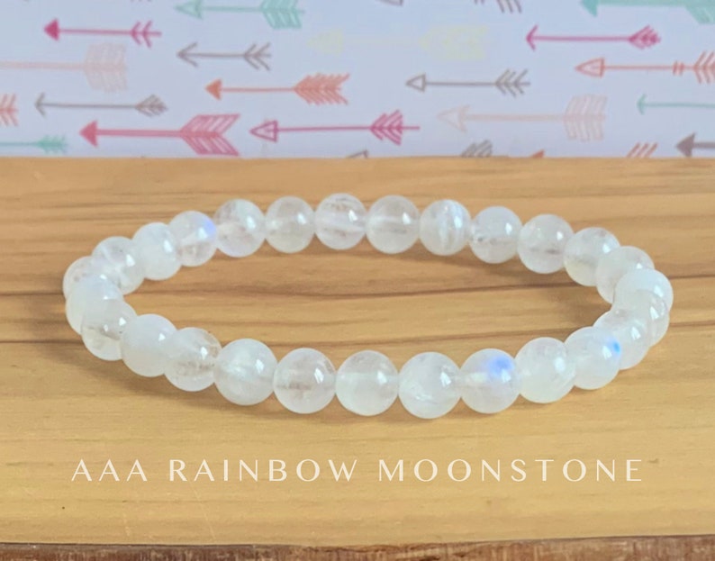 May include: A stretch bracelet composed of round, translucent white moonstone beads with subtle blue iridescence. The beads are strung in a single circular row. The text AAA RAINBOW MOONSTONE is printed in white capital letters below the bracelet.
