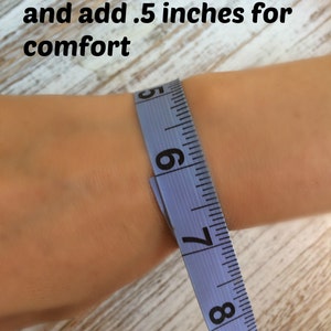 Puede incluir: Primer plano de una mu&ntilde;eca medida con una cinta m&eacute;trica azul. El texto dice: "Mide tu mu&ntilde;eca y a&ntilde;ade 1,3 cm para mayor comodidad". La escala est&aacute; en pulgadas.