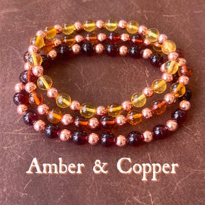 May include: Three beaded bracelets with amber and copper accents. The bracelets feature alternating rows of amber-coloured and dark red beads, interspersed with copper-coloured beads. The text "Amber & Copper" is at the bottom.