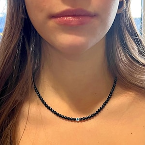 Può includere: Una collana di perline nere con un ciondolo a forma di occhio blu.