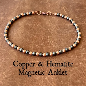 May include: Copper and hematite magnetic anklet. The anklet features small, round copper and dark gray beads strung together. The text "Copper & Hematite Magnetic Anklet" is printed on the brown surface beneath the anklet.