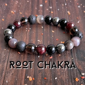 May include: A black beaded bracelet with red, grey, and black stones. The bracelet has the words "ROOT CHAKRA" written on the wooden surface it is resting on.