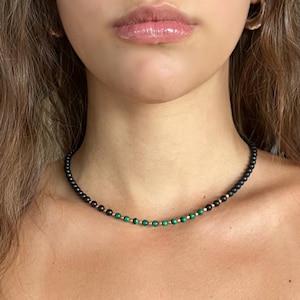 May include: A black beaded necklace with gold accents and green beads. The necklace is worn around the neck and is a choker style. The beads are small and round, creating a delicate and elegant look. The necklace is a piece of jewellery.